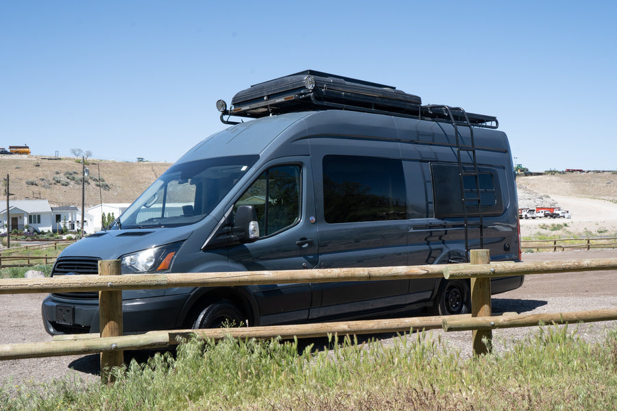 This dark gray Ford Transit camper van RV rental with rooftop tent and ladder is parked outdoors by a wooden fence.