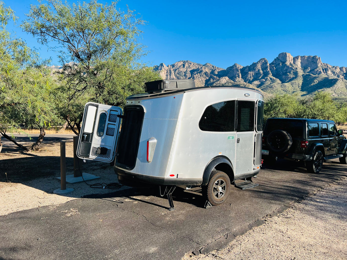Silver teardrop RV rental & black Jeep parked at a desert campsite with rugged mountains in the background.