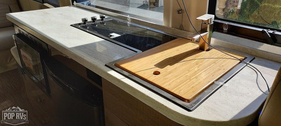 Modern RV kitchen counter with integrated cooktop, bamboo cutting board over the sink, and microwave. Perfect for an rv rental.