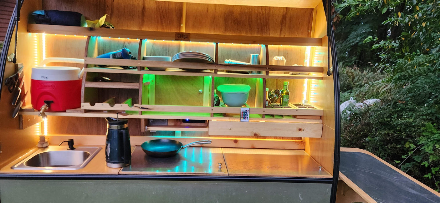 Bright, organized camper kitchen setup, ideal for an RV rental. Features shelves, LED lights, sink, cooler, and cooking pan.