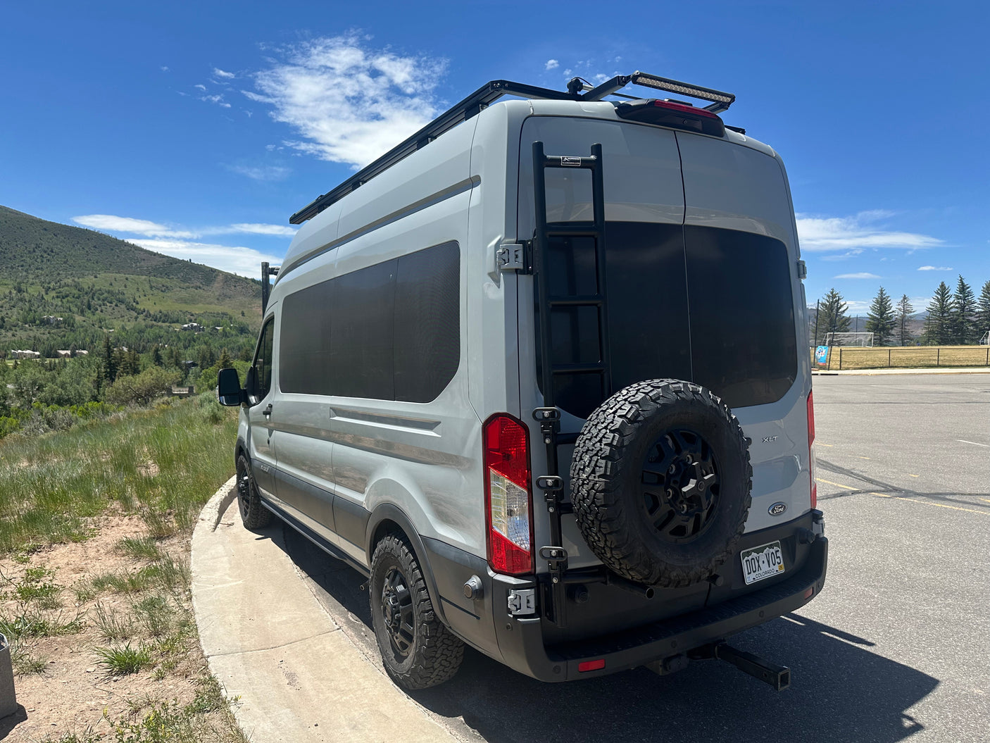 Adventure-ready gray Ford Transit camper van with a roof rack, ladder, and spare tire, ideal for an RV rental trip.