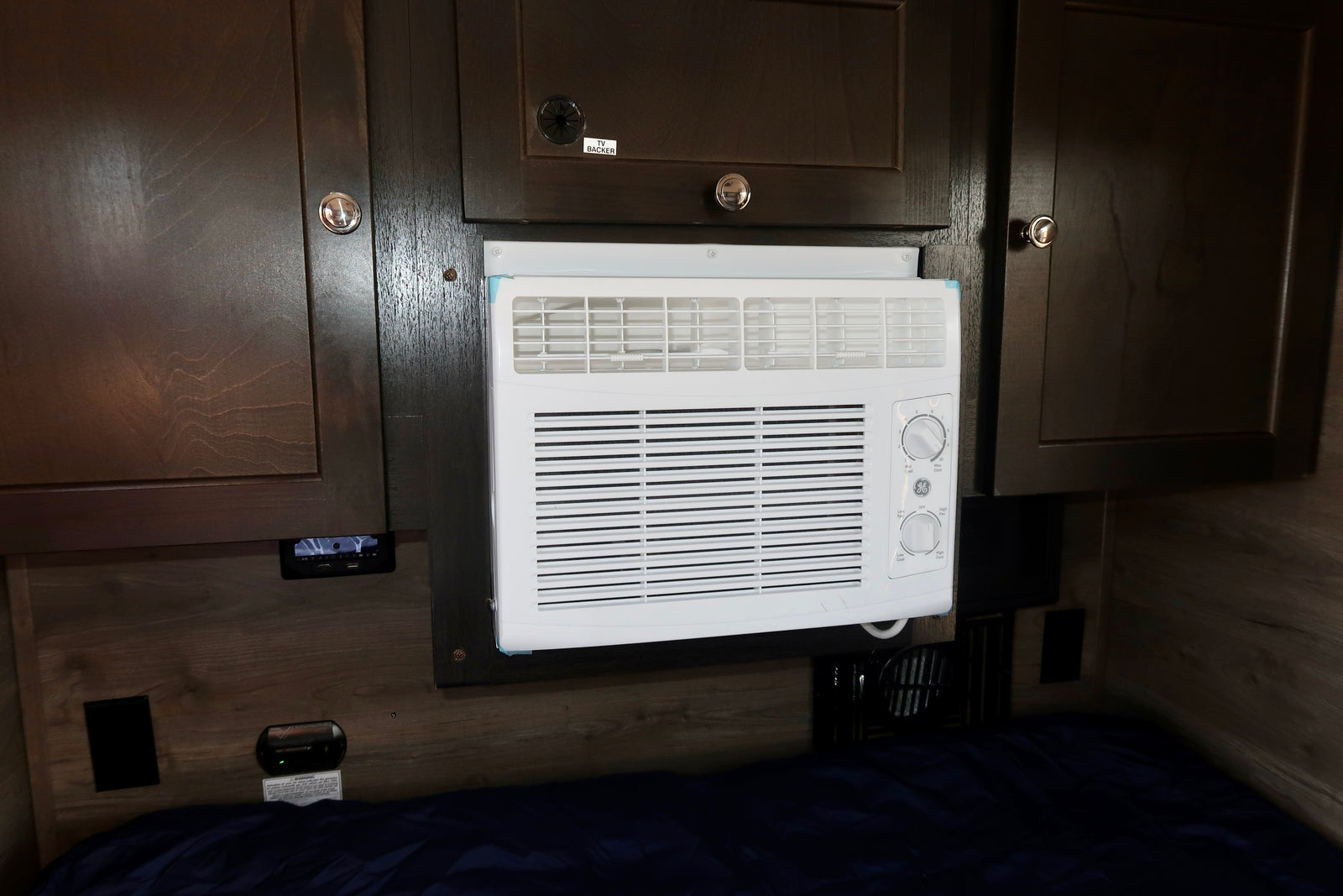 Compact AC unit in an RV rental, integrated into dark wood cabinetry above a bed.