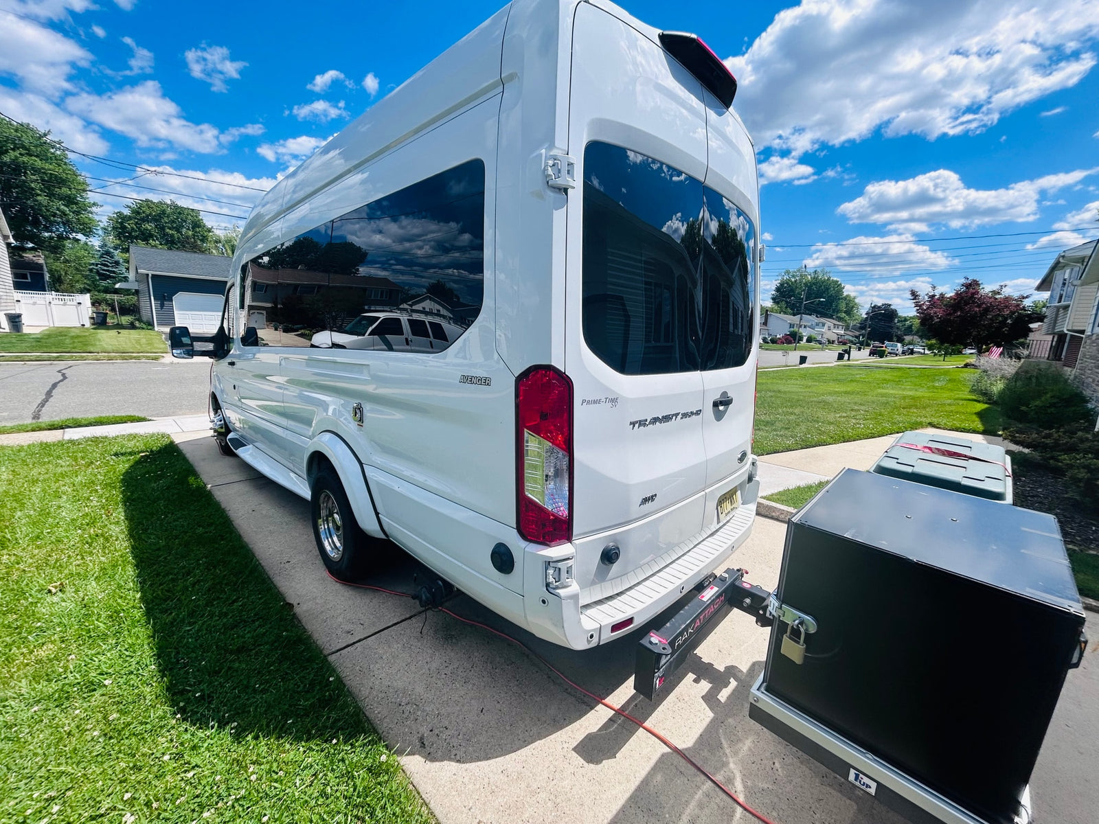 A white converted Ford Transit van, ideal for RV rental, with a black cargo carrier on its rear hitch. Parked on a sunny suburban driveway.