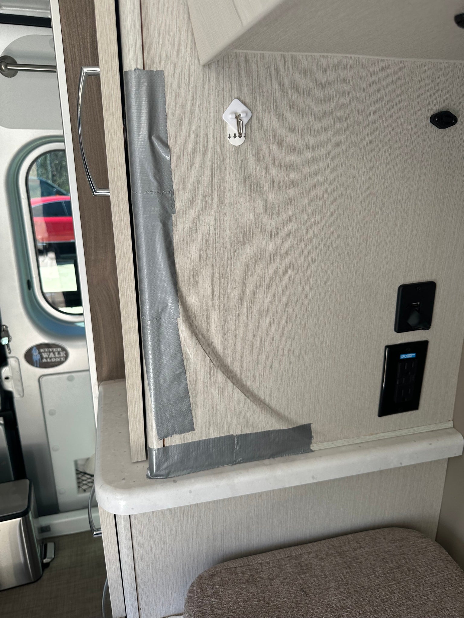 Gray duct tape temporarily secures a cabinet/door in an RV rental. Features a wall hook, USB/GFI outlets, and a "Never Walk Alone" sticker.