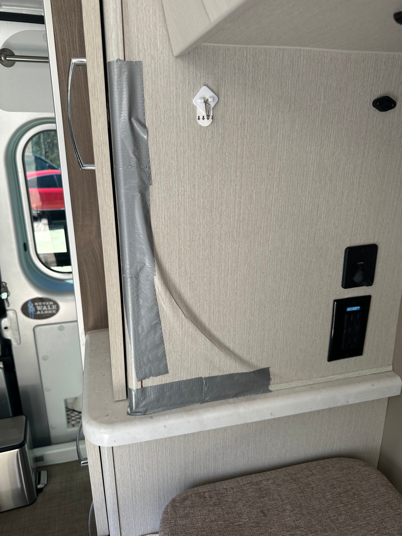 Gray duct tape temporarily secures a cabinet/door in an RV rental. Features a wall hook, USB/GFI outlets, and a "Never Walk Alone" sticker.
