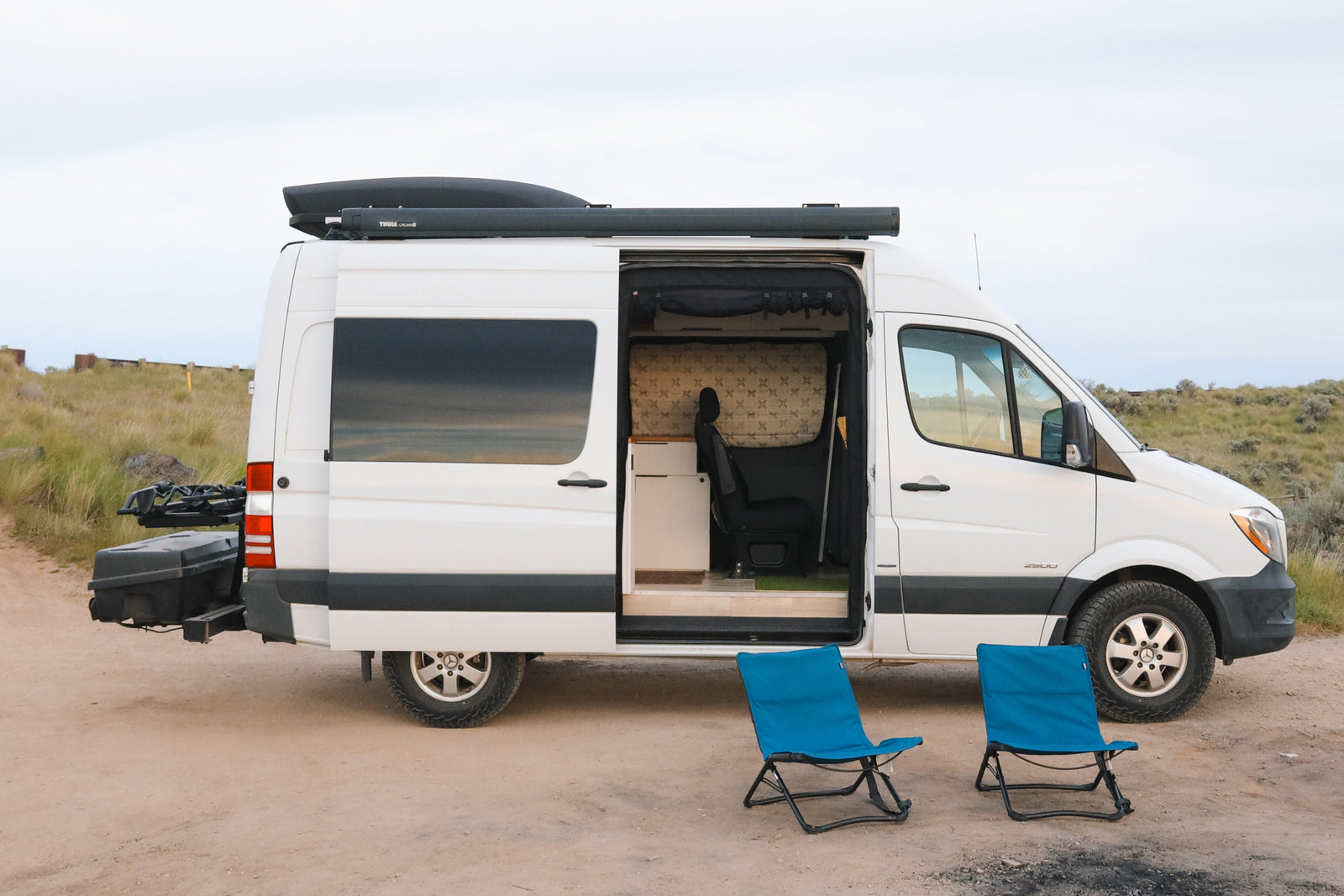 White RV rental Sprinter camper van with open door and interior view. Two blue chairs await a camping adventure.