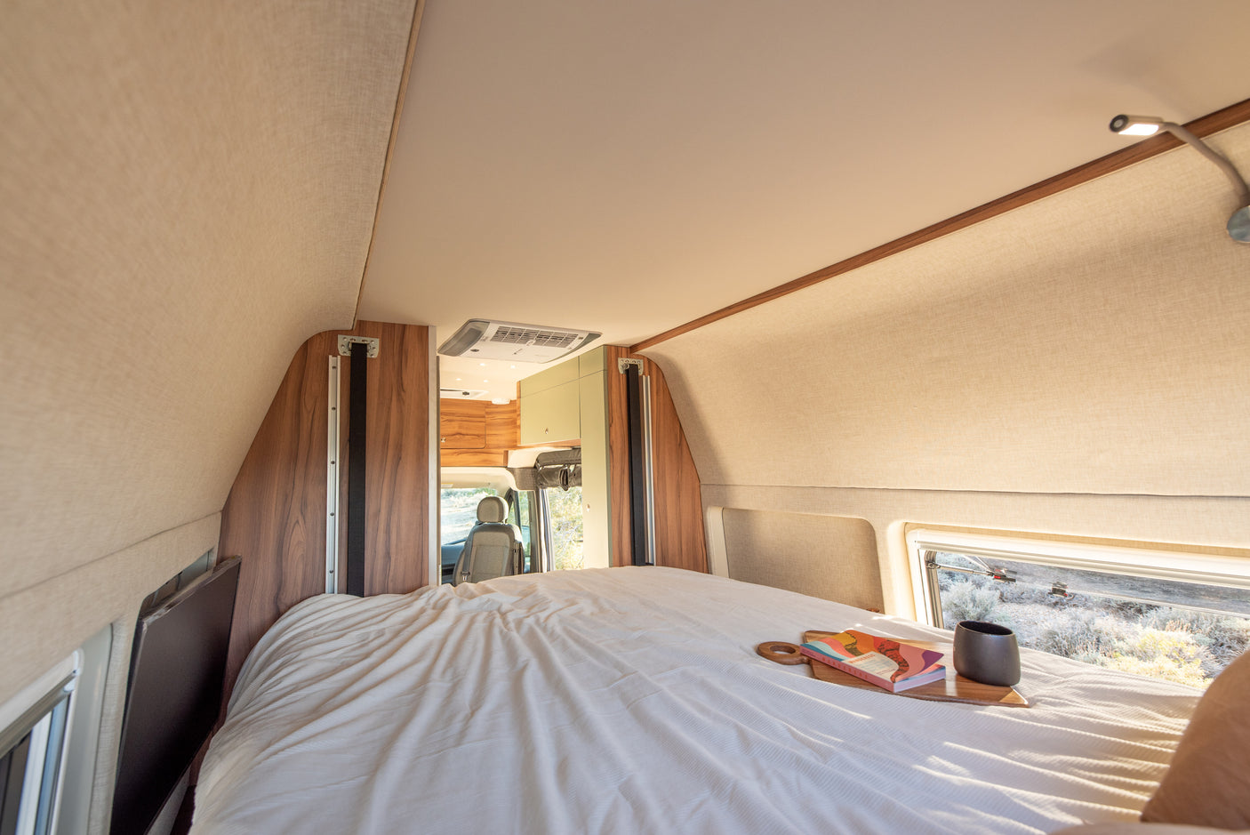 Modern RV rental bedroom: a cozy bed with a book, mug, and a window revealing a scenic outdoor view.