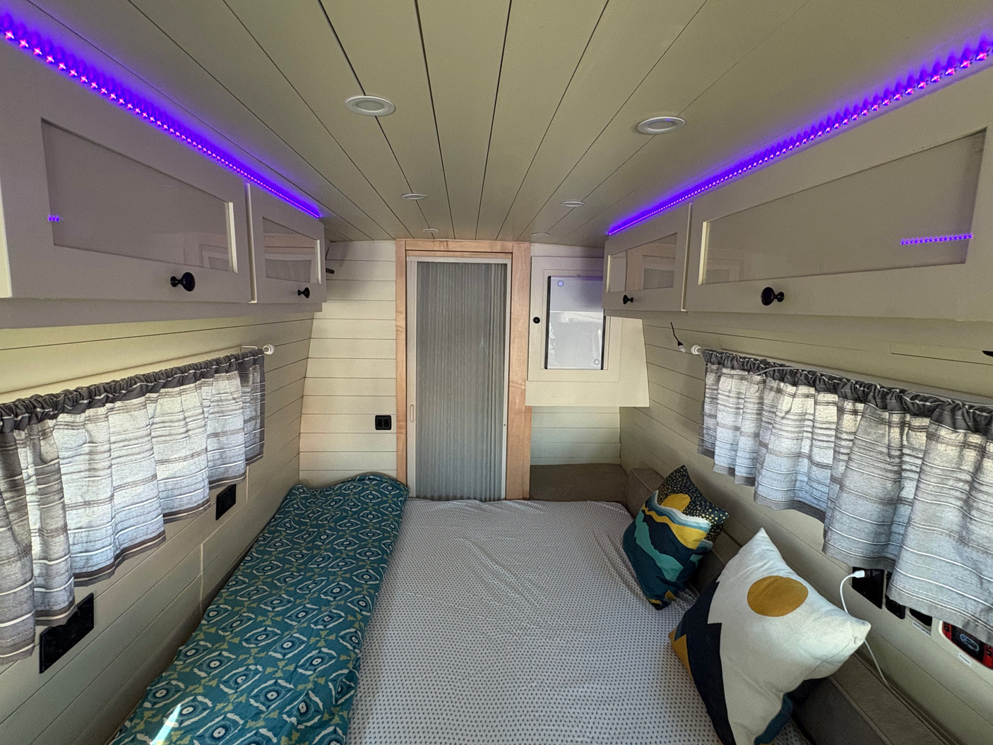 Cozy RV rental interior with a comfortable bed, vibrant purple LED lighting, overhead cabinets, and window curtains.