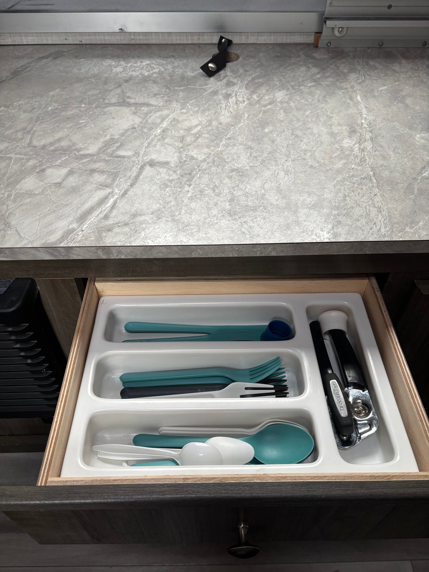 Open RV rental kitchen drawer featuring an organizer with teal, white, and black plastic cutlery and a can opener.