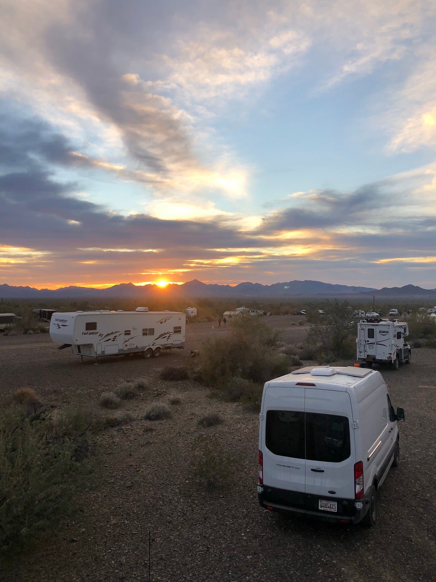 Desert RV camping at sunset. Vans & RVs, including a potential RV rental, are parked. Mountains and a colorful sky complete the serene scene.