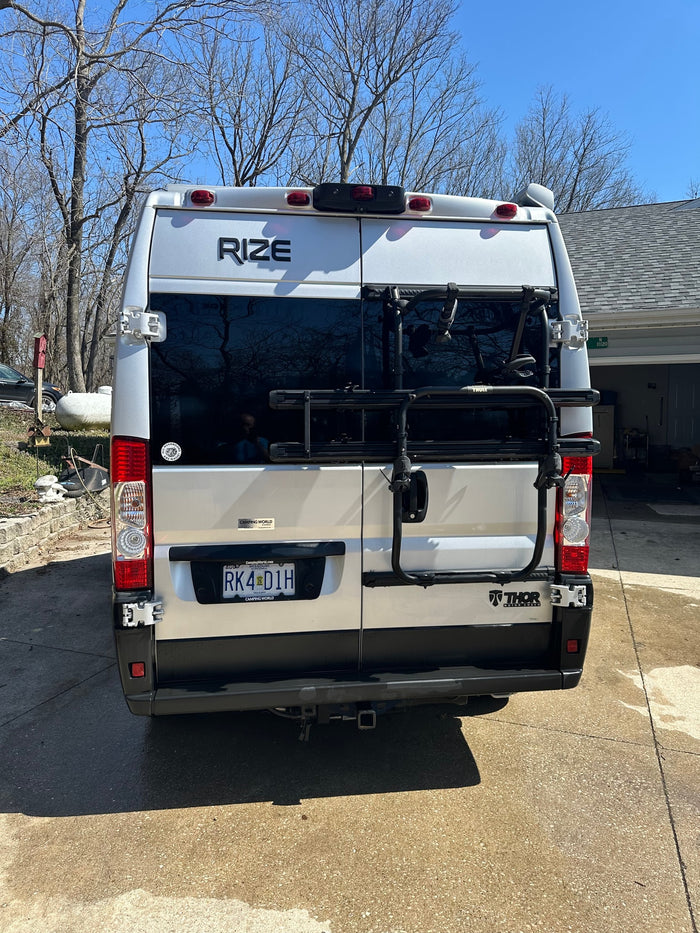 Silver **RV rental** camper van with a rear bike rack and hitch, parked outdoors. Features 'Rize' branding.