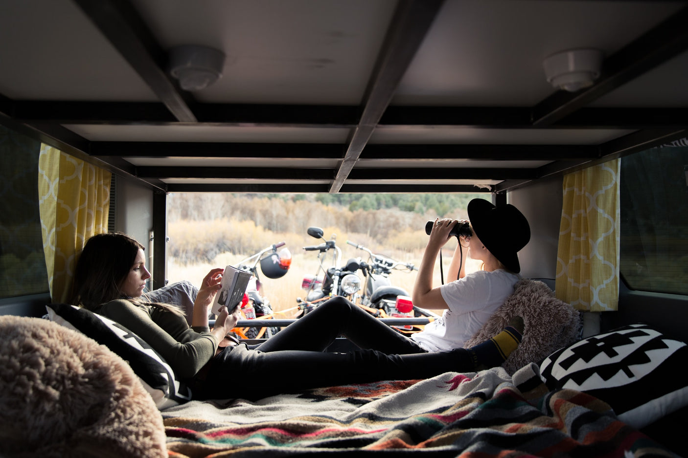 Relaxing inside an RV rental, two women enjoy downtime: one reading, the other observing the scenic view with binoculars, motorcycles outside.