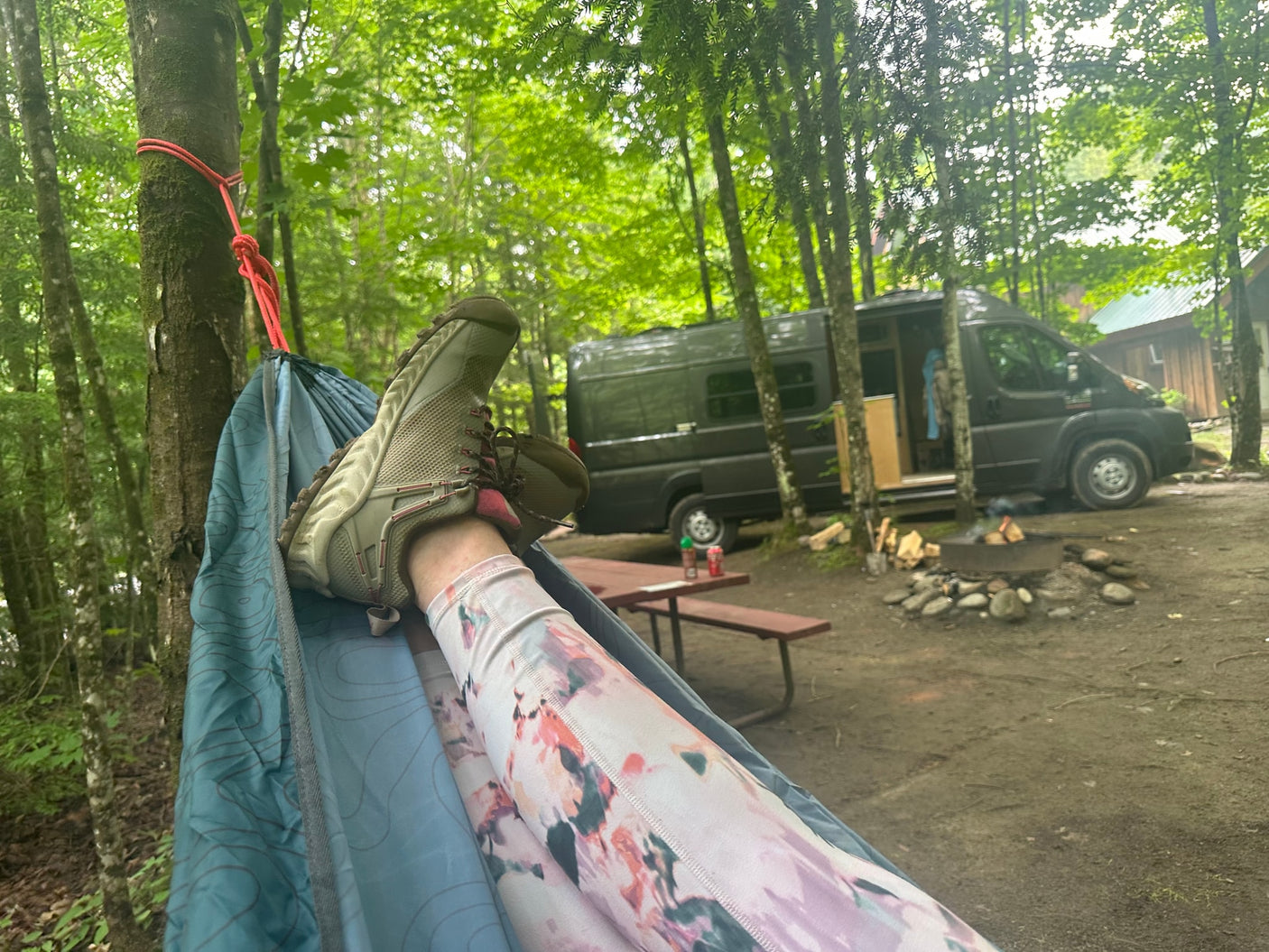Legs in a hammock at a serene RV rental campsite. A dark camper van, picnic table, and fire pit are visible.