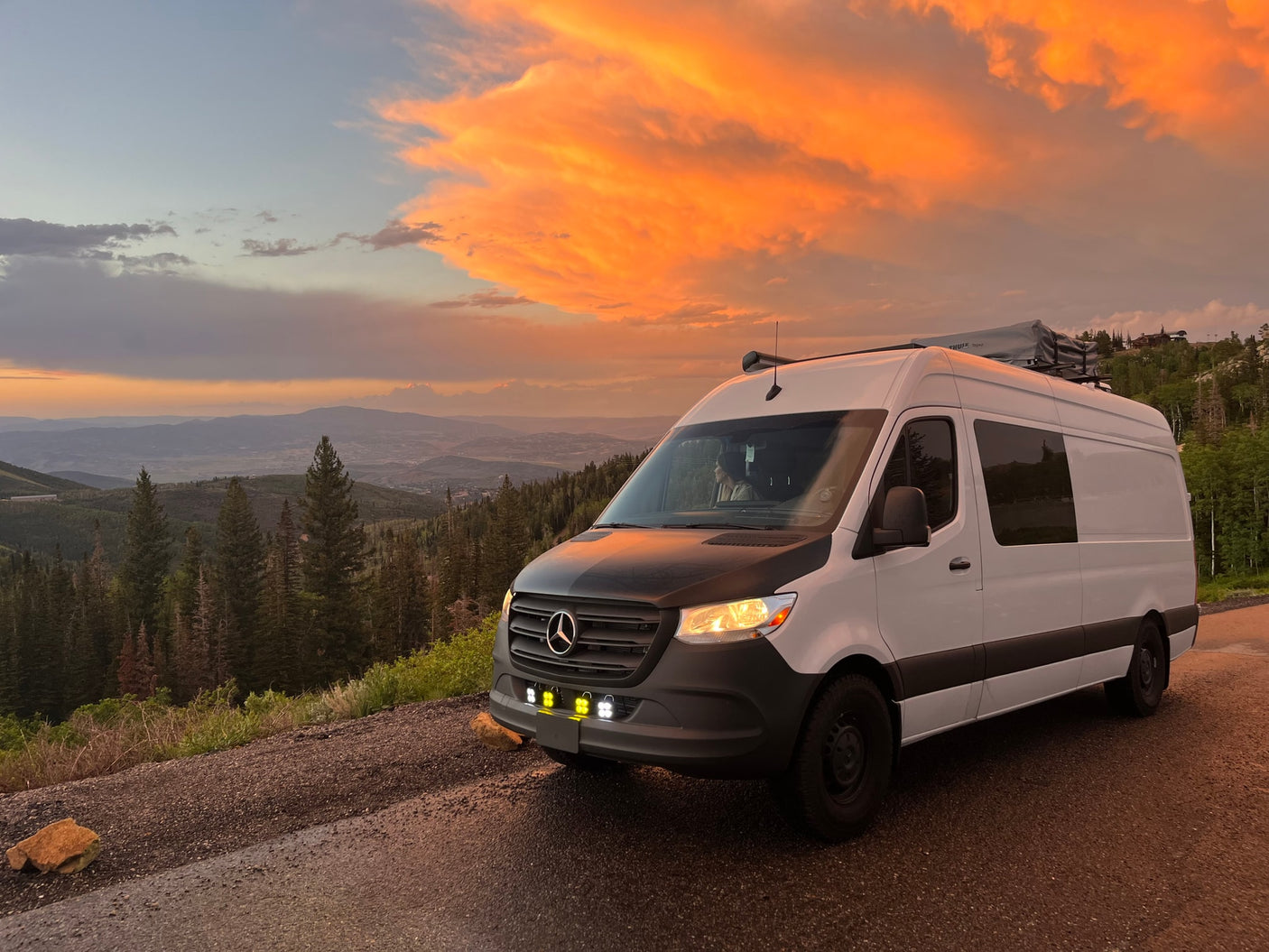A white RV rental van overlooking a sunset-lit valley. Vibrant orange clouds, mountains, and forest. Ready for adventure.