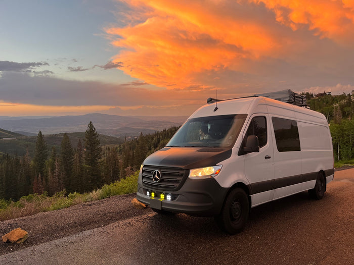 A white RV rental van overlooking a sunset-lit valley. Vibrant orange clouds, mountains, and forest. Ready for adventure.