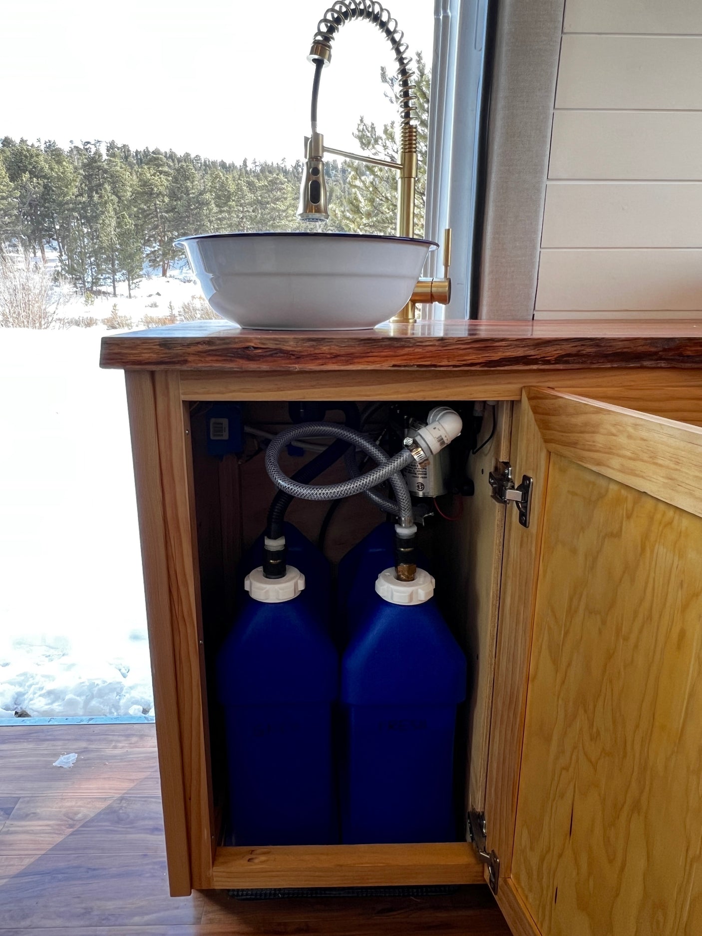 RV rental kitchen: stylish sink, snowy forest view. Underneath, a practical off-grid water system with two blue tanks.