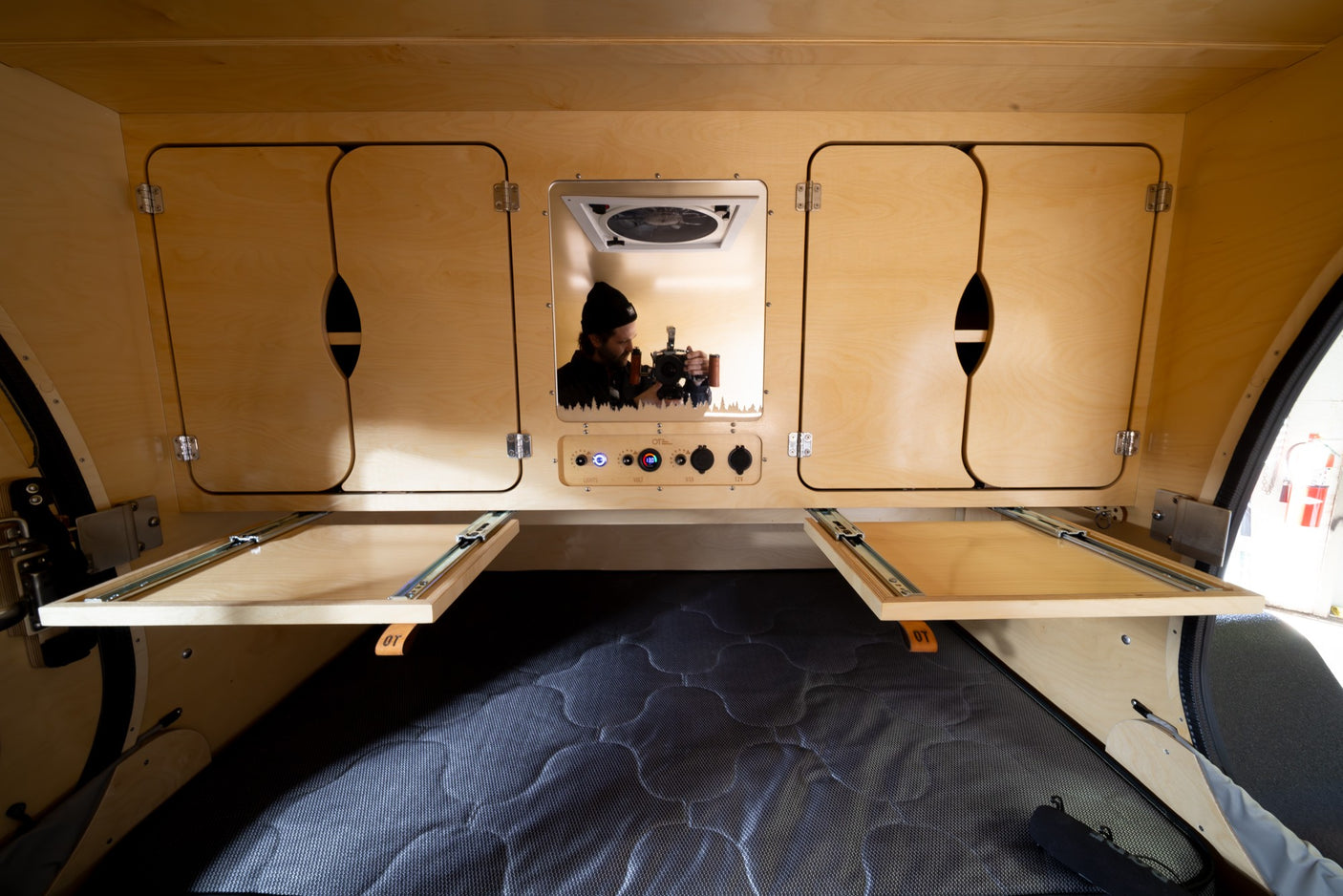 Teardrop RV rental interior: wooden cabinets, control panel with 12V/USB, pull-out tables, and photographer's mirror reflection.