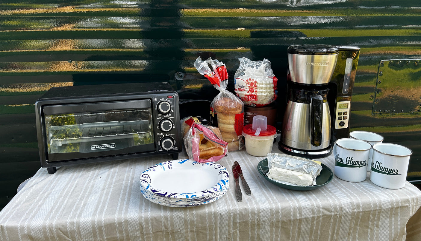 Outdoor breakfast setup by an RV rental: toaster oven, coffee maker, bagels, and 'Glamper' mugs on a striped tablecloth.