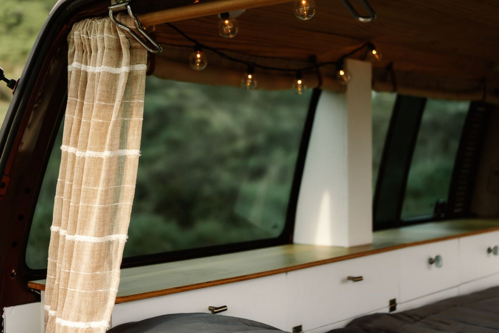 Inside a cozy RV rental: warm string lights, a light curtain, wood details, and a large window overlooking green nature.
