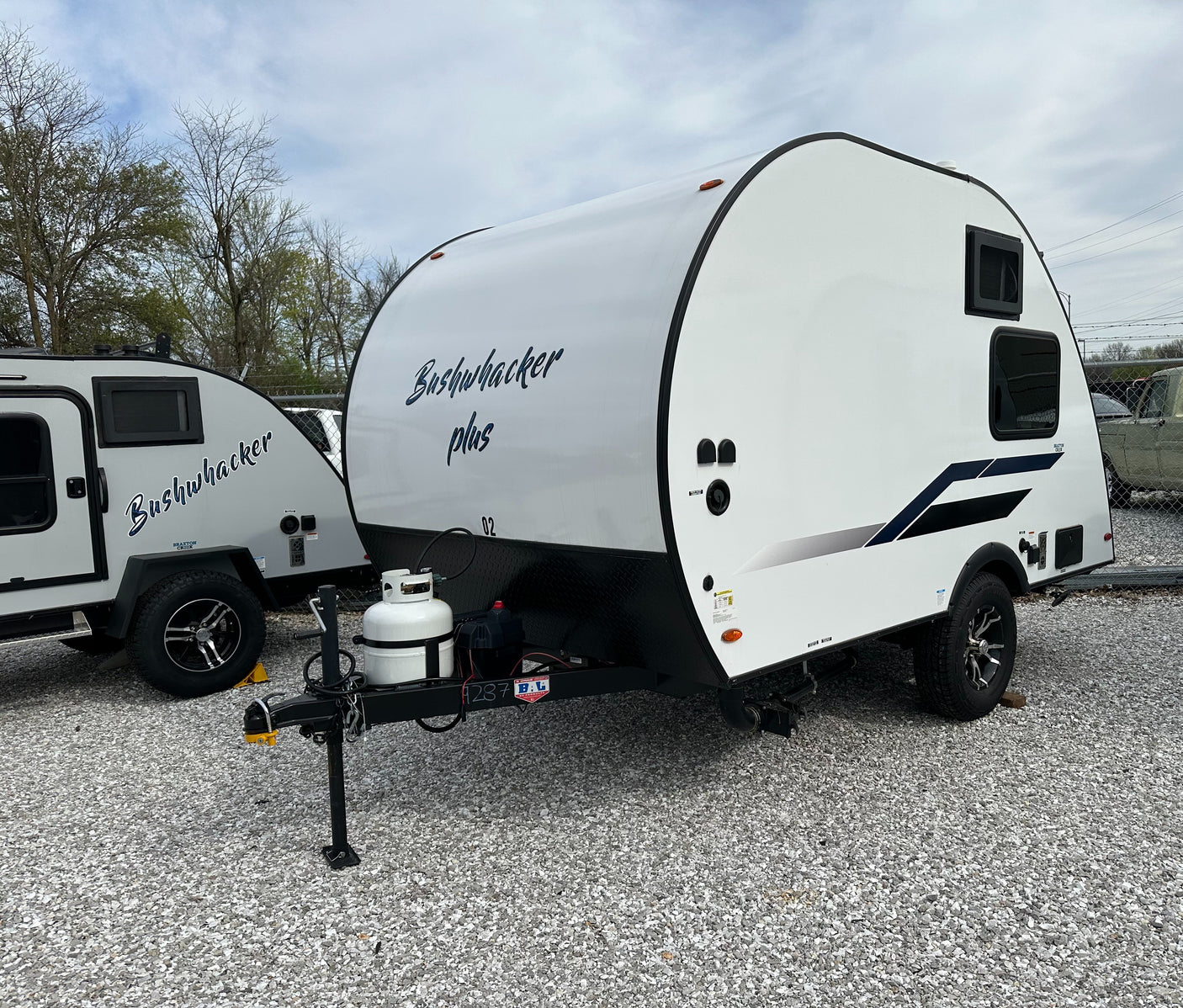 Two Bushwhacker travel trailers, a white "Plus" model in front, are parked on gravel, ideal for an RV rental adventure.