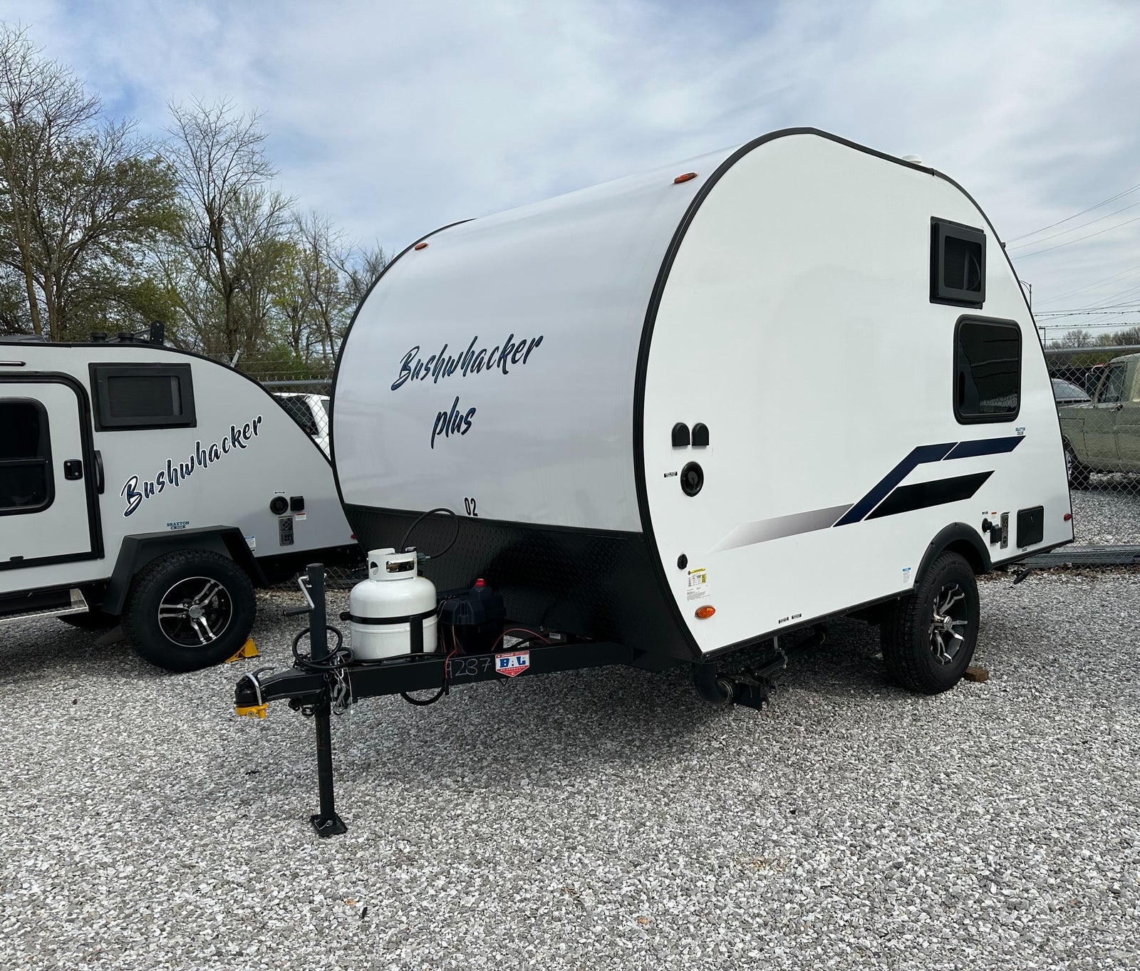 Two Bushwhacker travel trailers, a white "Plus" model in front, are parked on gravel, ideal for an RV rental adventure.