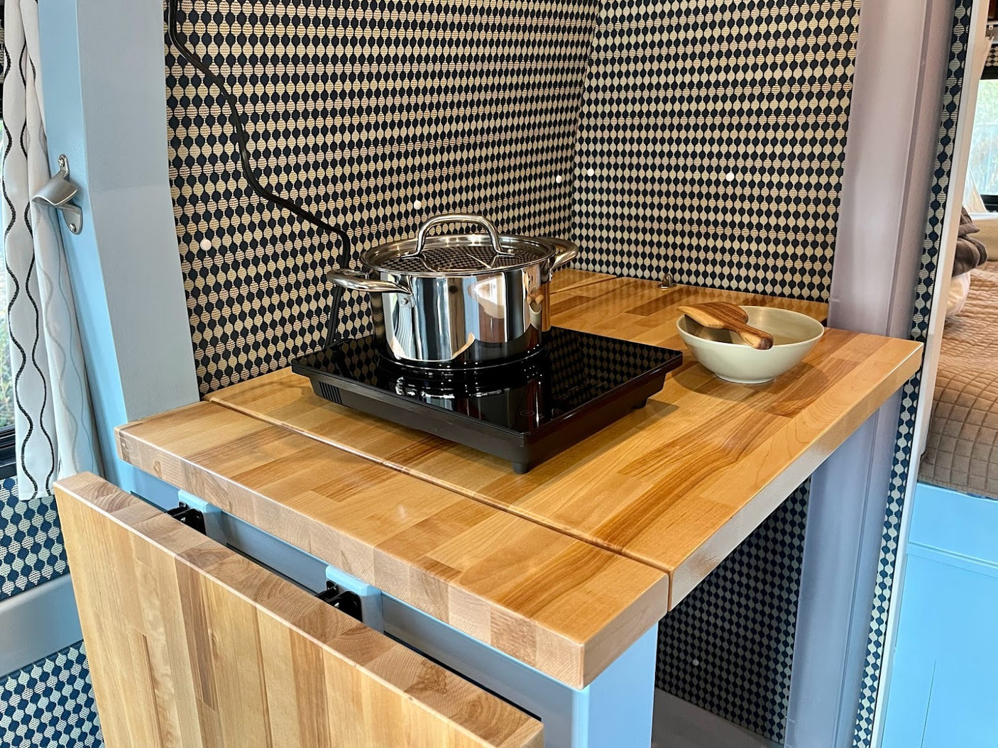 This RV rental kitchen features a light wood counter, induction cooktop with a pot, and a stylish patterned wall.