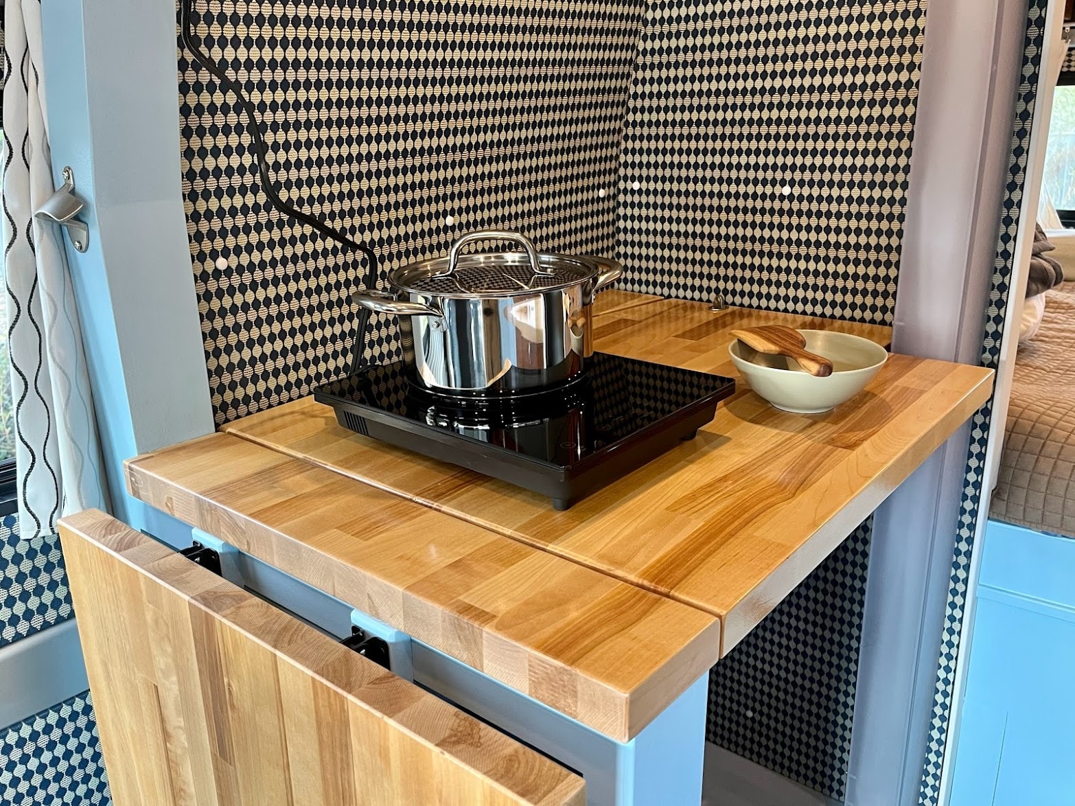 This RV rental kitchen features a light wood counter, induction cooktop with a pot, and a stylish patterned wall.