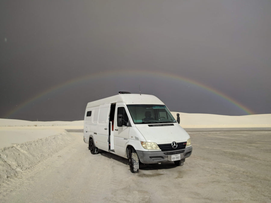 RV rental camper van parked on White Sands' gypsum with a rare rainbow arching across a dramatic stormy sky.