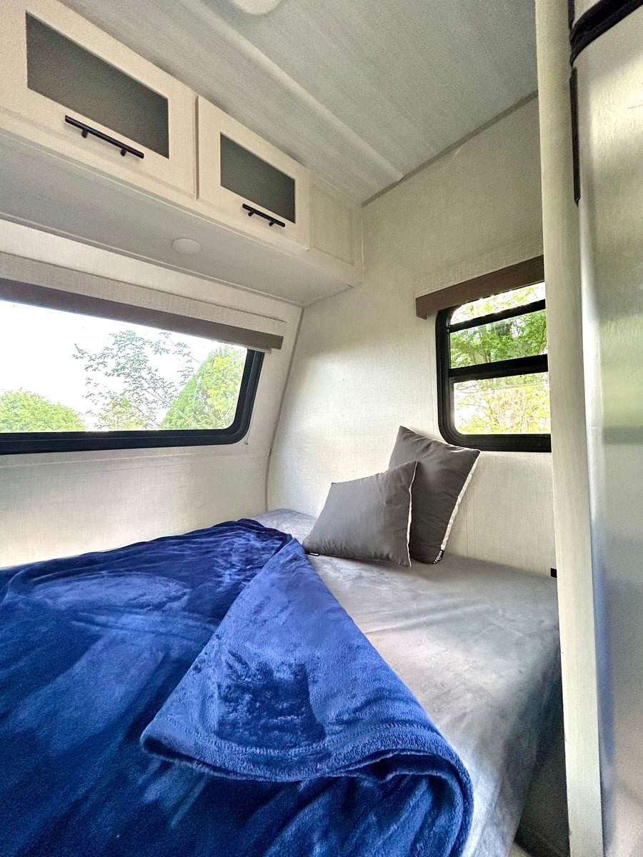 Enjoy this RV rental's cozy bedroom: a bed with a soft blue blanket, gray pillows, and sunny windows overlooking trees.