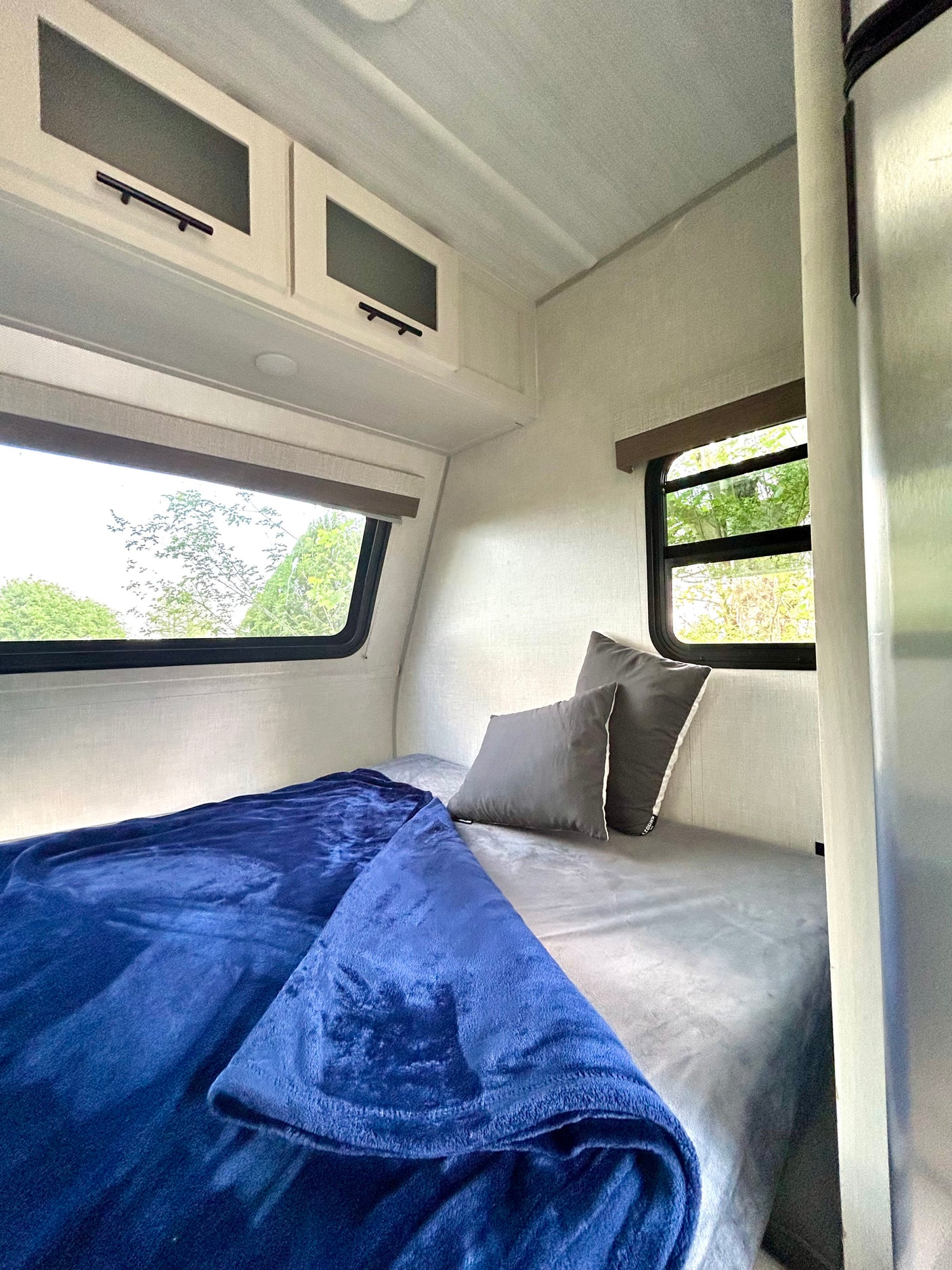 Enjoy this RV rental's cozy bedroom: a bed with a soft blue blanket, gray pillows, and sunny windows overlooking trees.