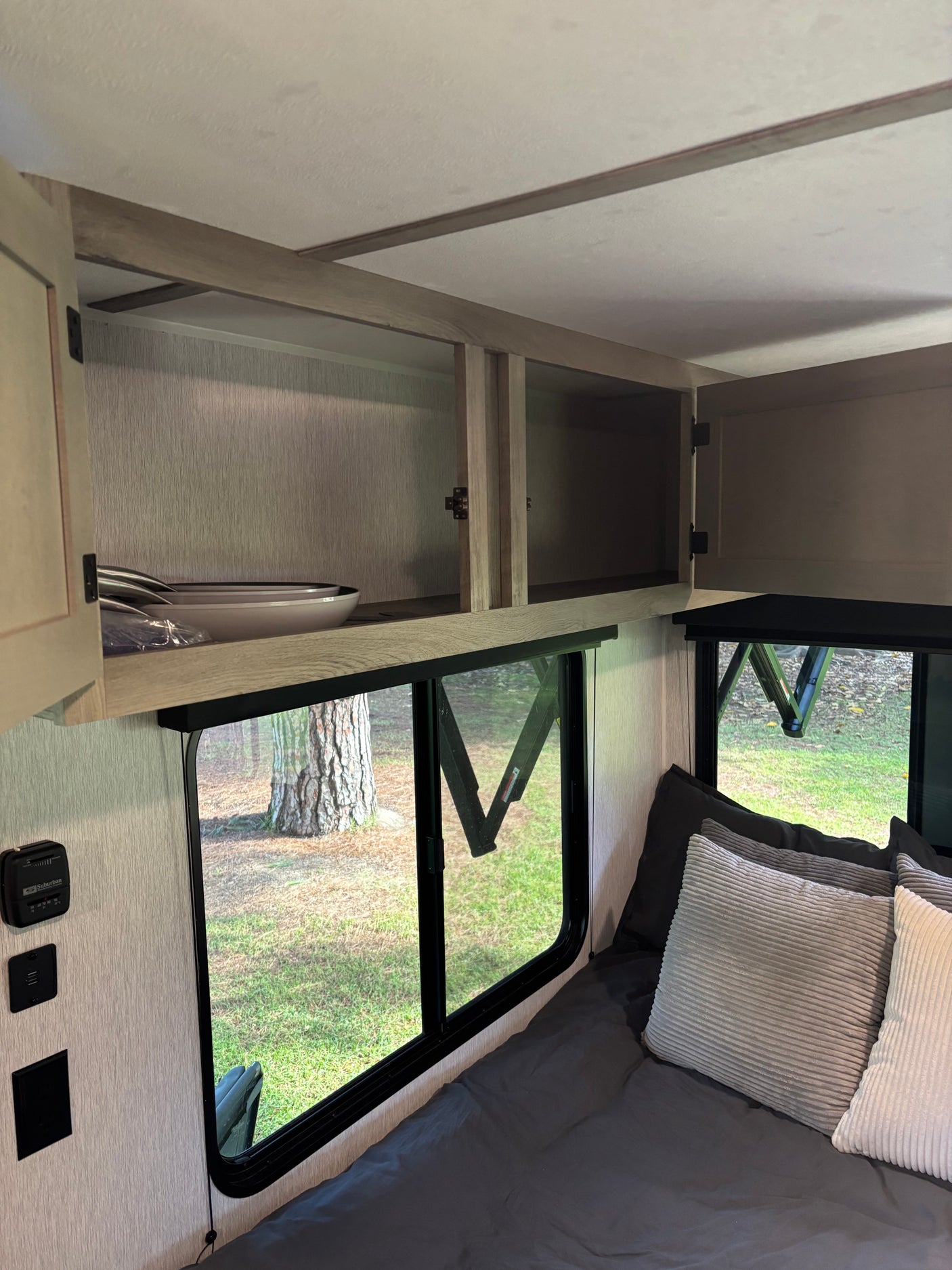 RV rental interior: open overhead cabinets with dishes, large windows overlooking trees, a thermostat, and a made-up bed.