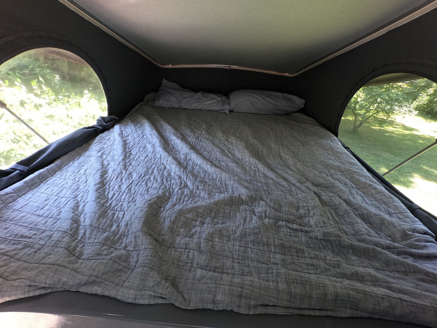 Inside an RV rental pop-top, a comfortable bed with grey bedding and pillows offers views of nature.