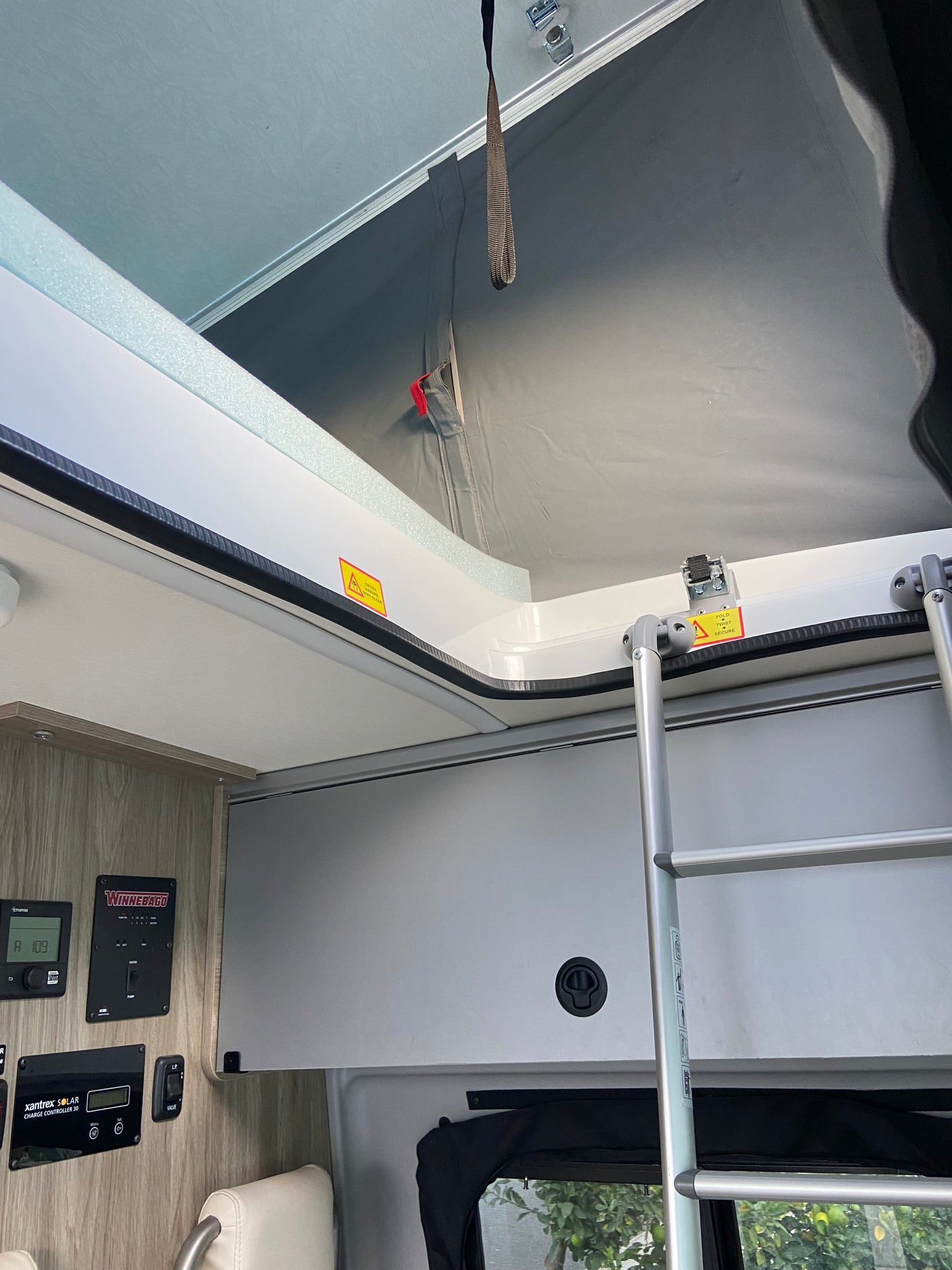 Inside an RV rental: a pop-top roof access with ladder, warning labels (crush hazard, secure), and control panels (Winnebago, solar).