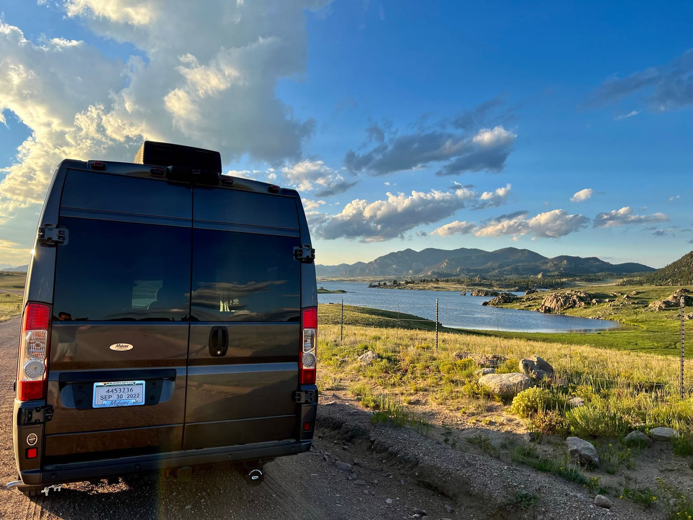A dark RV rental overlooks a scenic lake with mountains under a cloudy blue sky. Perfect for a nature road trip adventure!