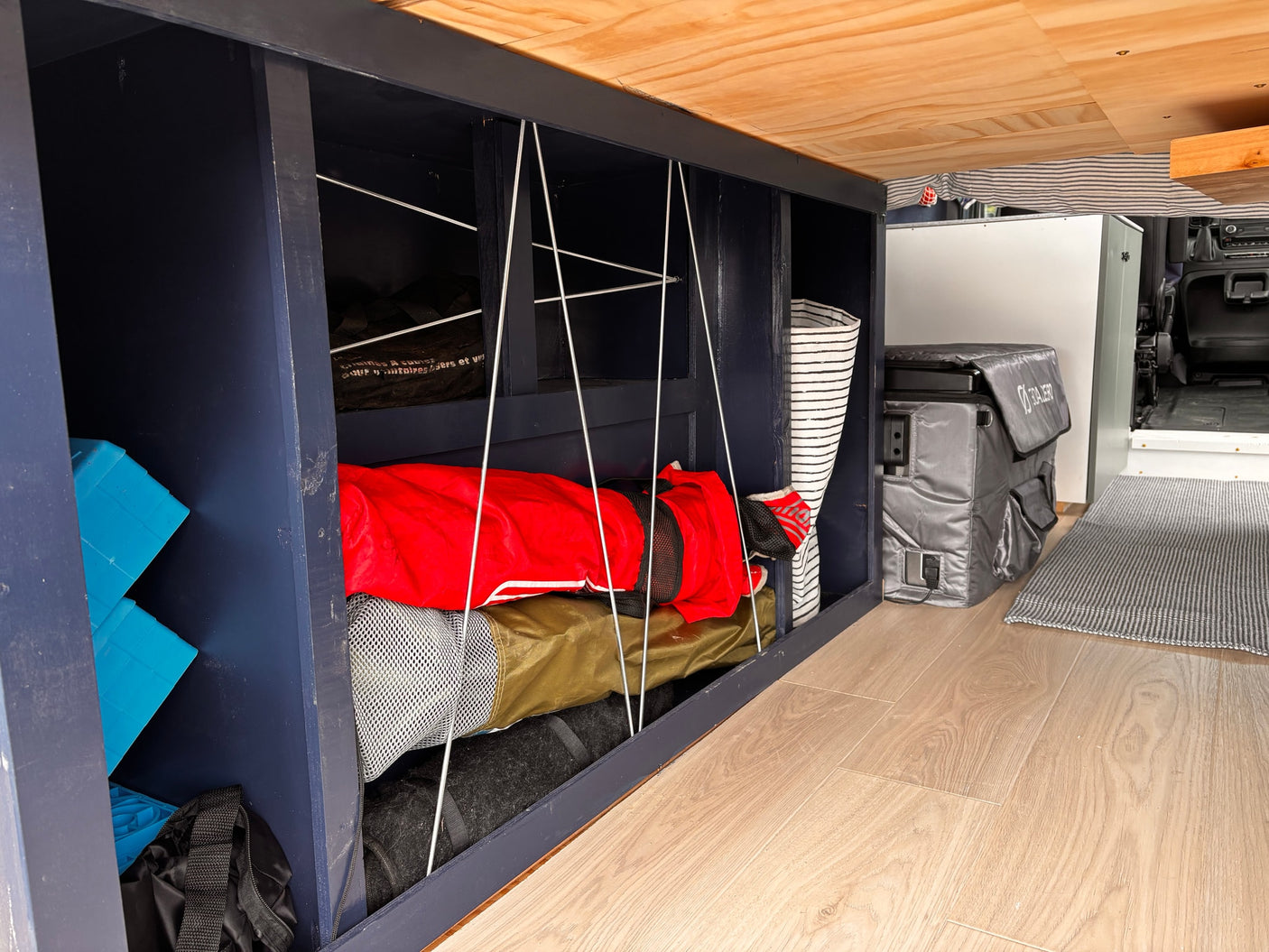 Custom RV rental storage: dark blue shelves with bungee cords organize camping gear, blue blocks, and a cooler on light wood floors.