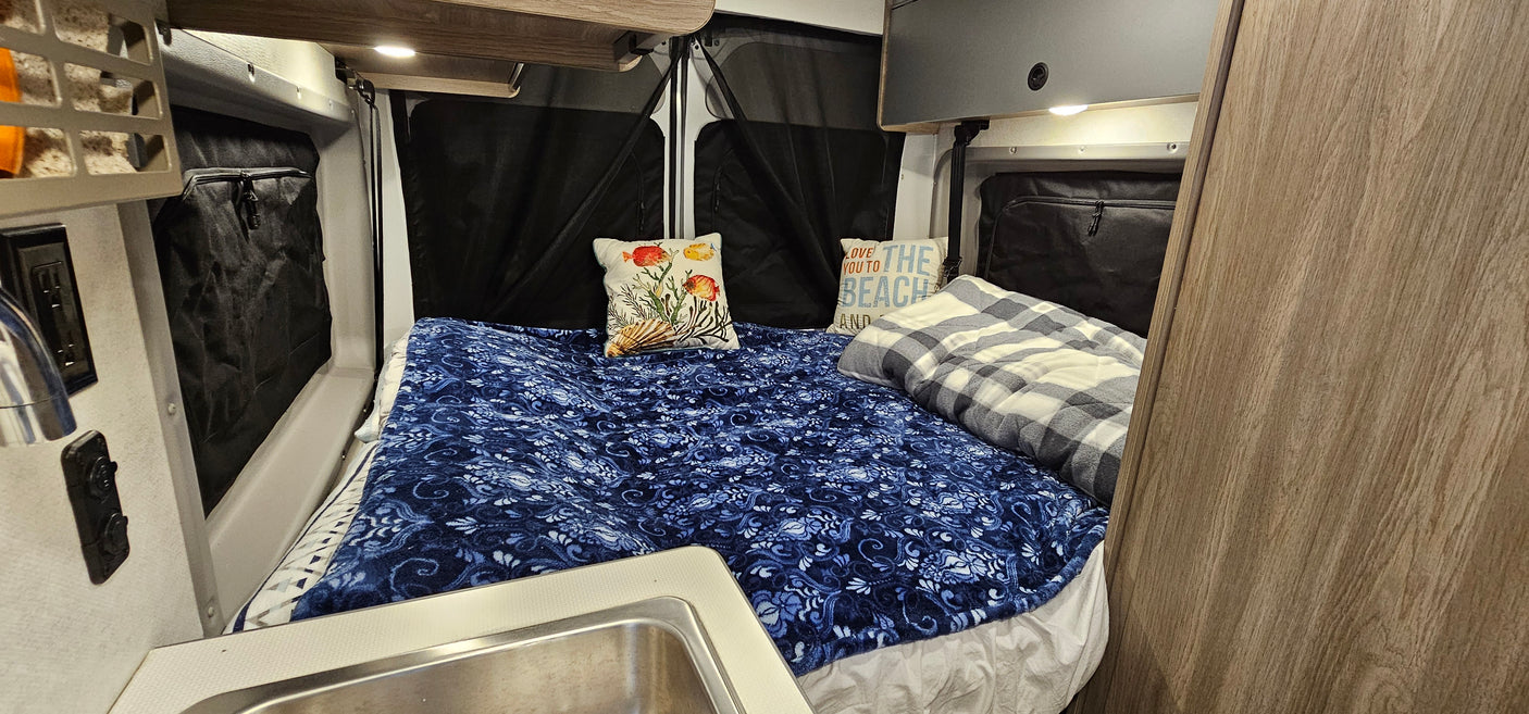 Experience an RV rental: this cozy camper interior features a bed with blue bedding, pillows, and a small sink.