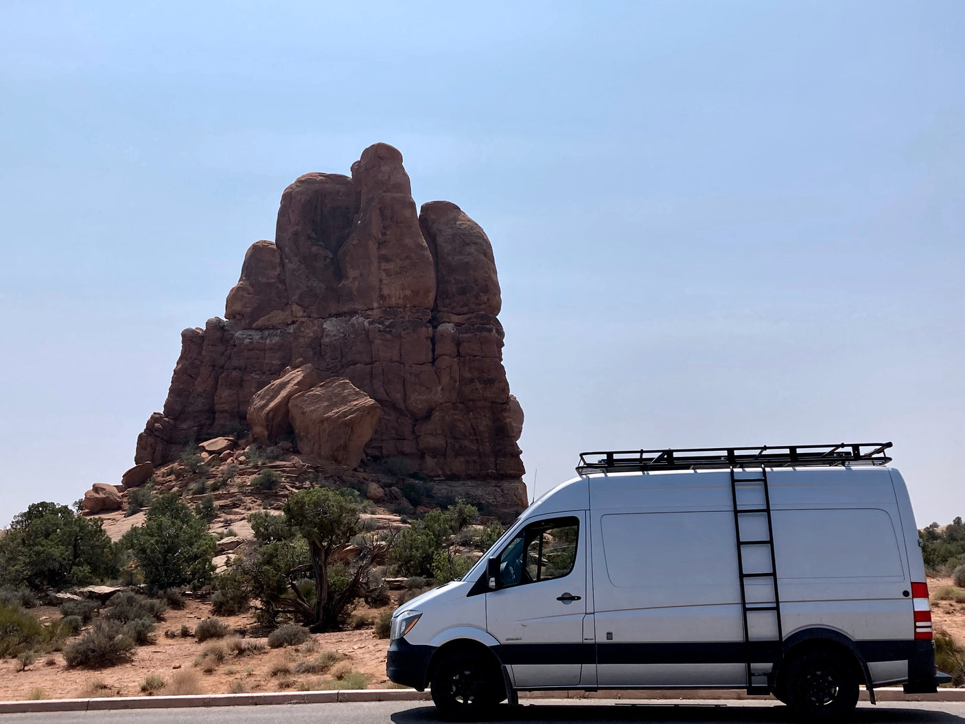 A white Sprinter-style **rv rental** van with a roof rack and ladder parked next to large red rock formations in a sun-drenched desert.