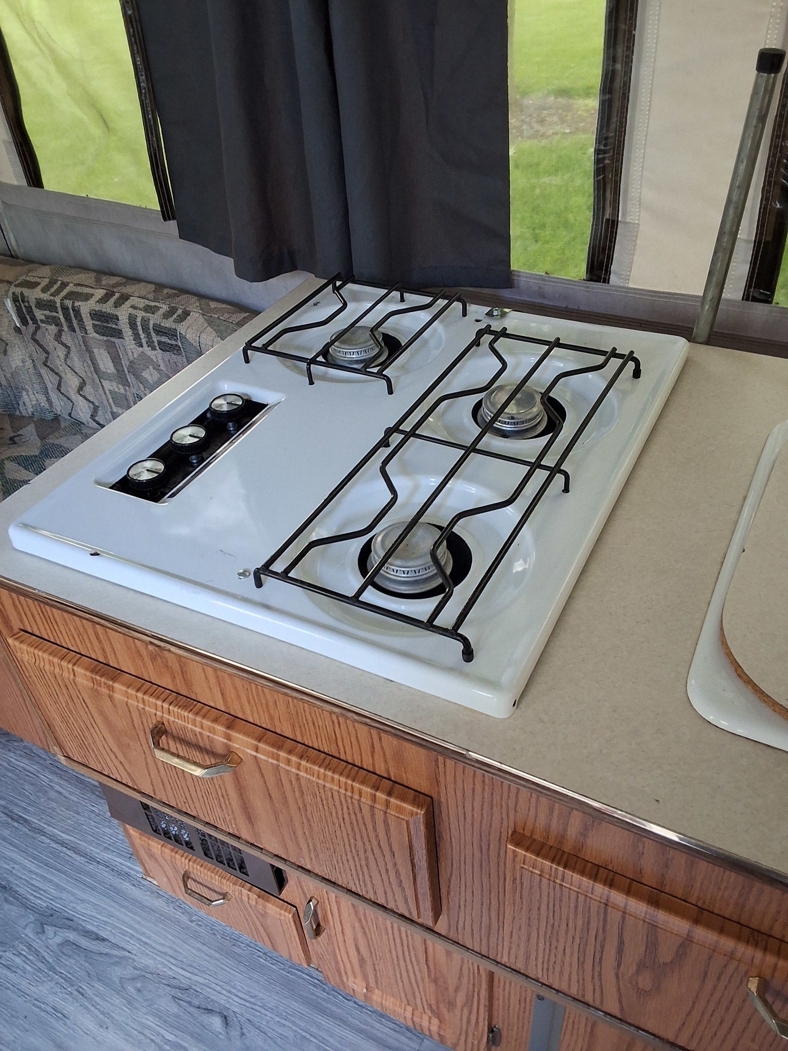 An RV rental kitchen showcasing a clean, white three-burner gas stovetop, wood cabinetry, and a bright window view.