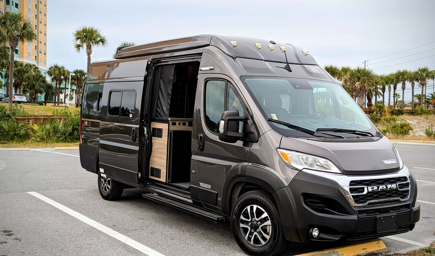 A stylish gray Winnebago Solis camper van, ideal for RV rental adventures. Side door open, parked by coastal palm trees.