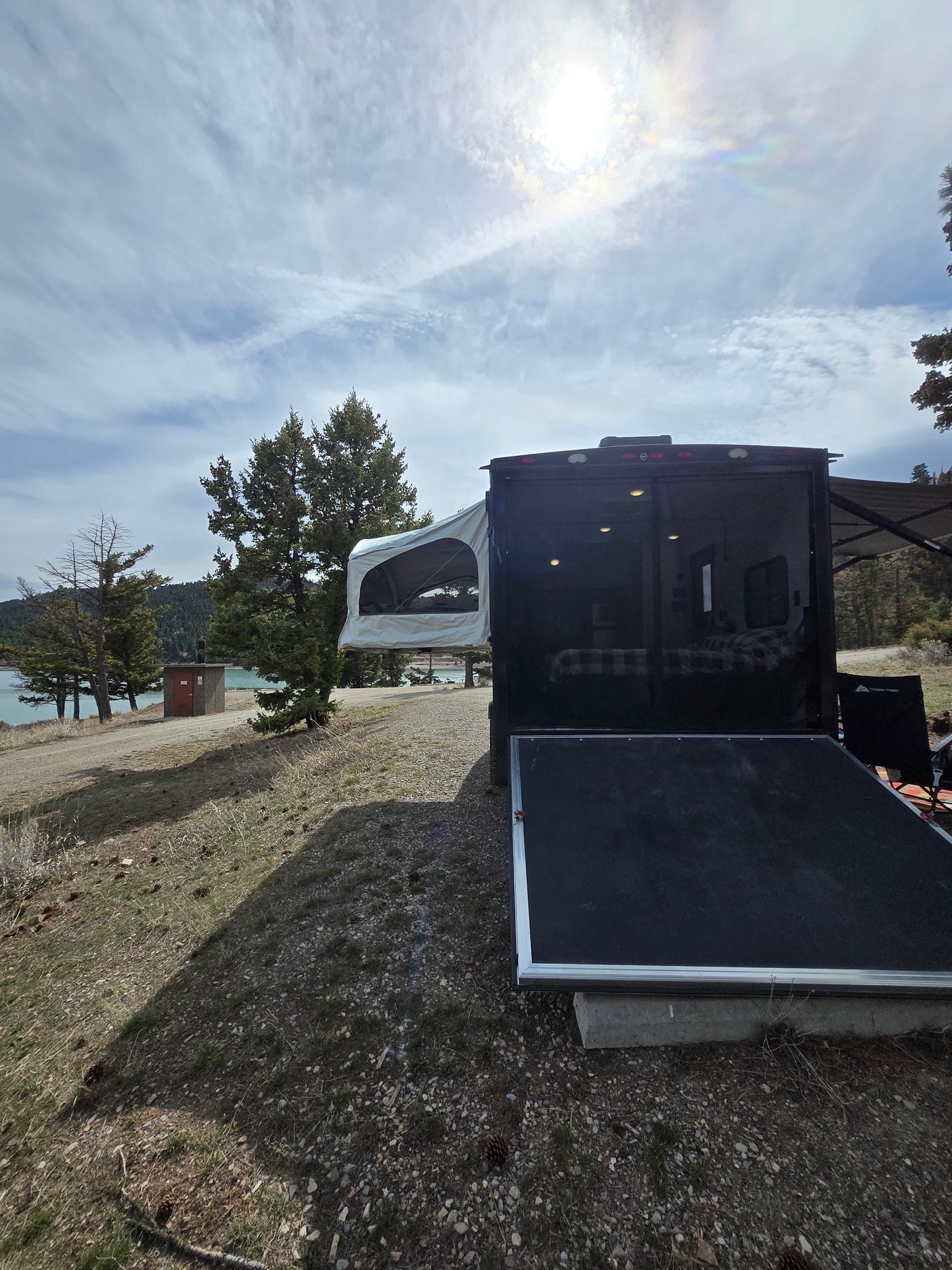 Scenic rv rental at a lakeside campsite, featuring a toy hauler with open ramp and pop-out tent. Bright sun overhead.