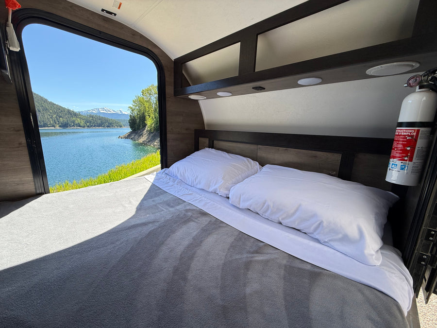 Inside an RV rental, a cozy bed faces a large window with a beautiful view of a lake and snow-capped mountains.