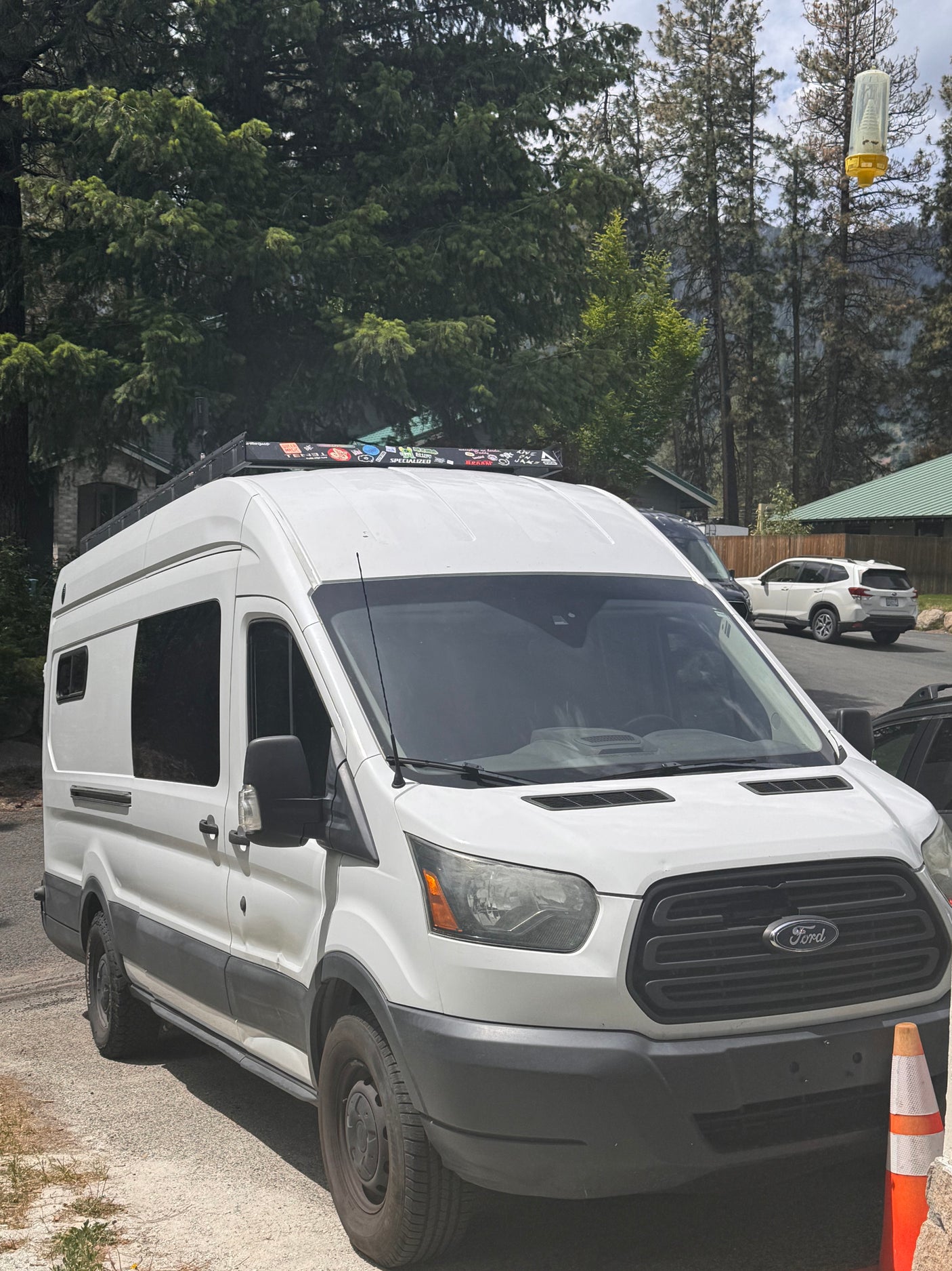 This white Ford Transit camper van, an ideal RV rental, is parked outdoors among tall trees, ready for adventure.
