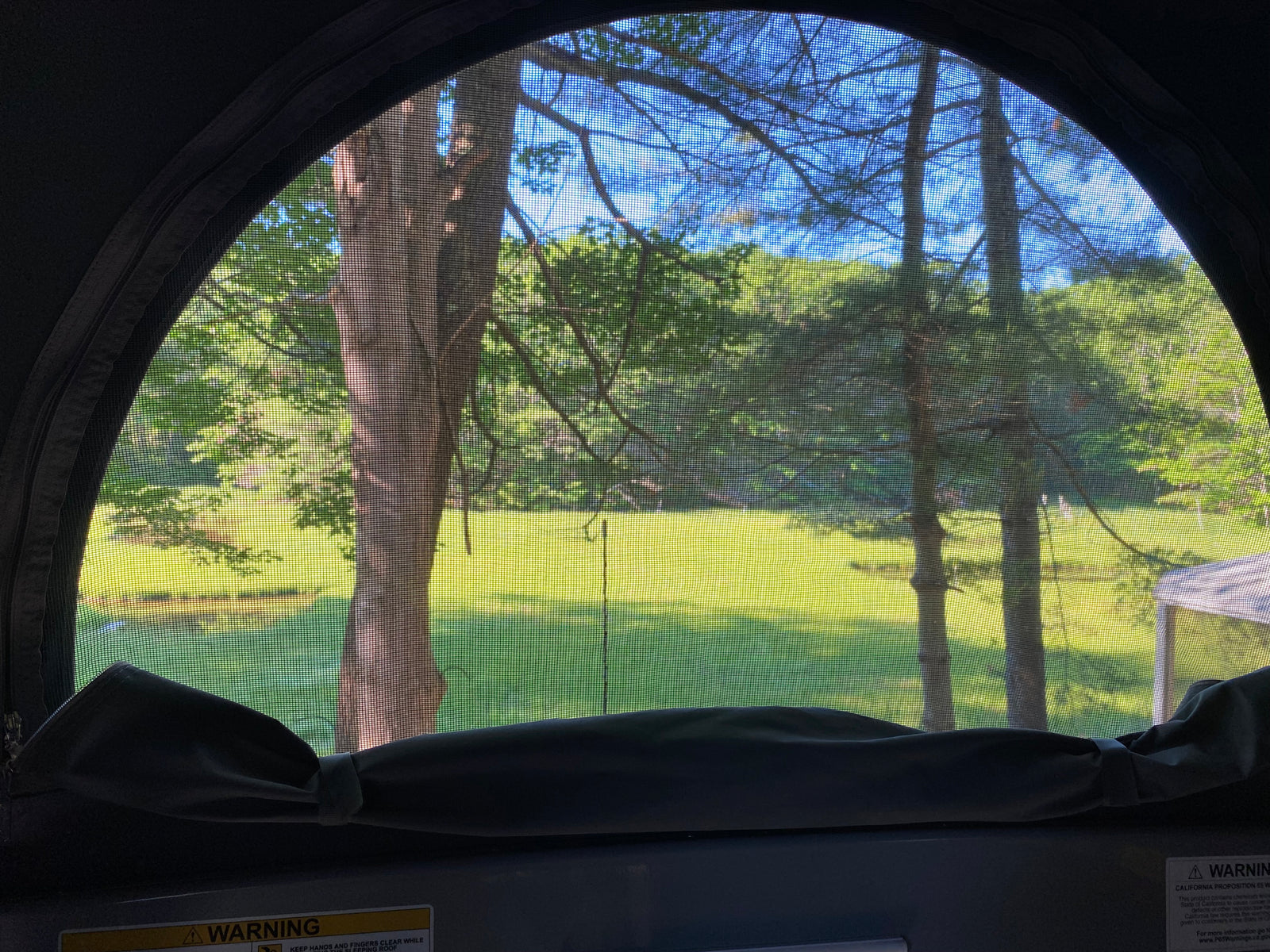 Looking out an RV rental's arched screened window reveals a vibrant green field, trees, and a clear blue sky.