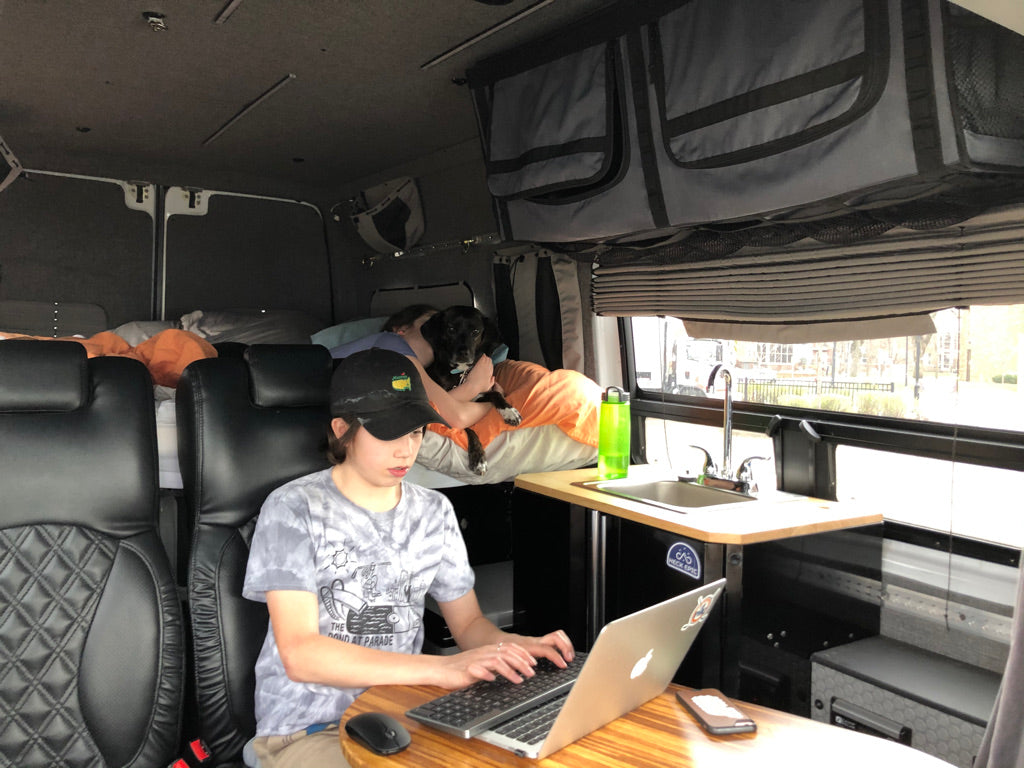 Inside an **RV rental**, a person types on a laptop at a wooden table while a dog rests on a bed. Features a compact kitchen.