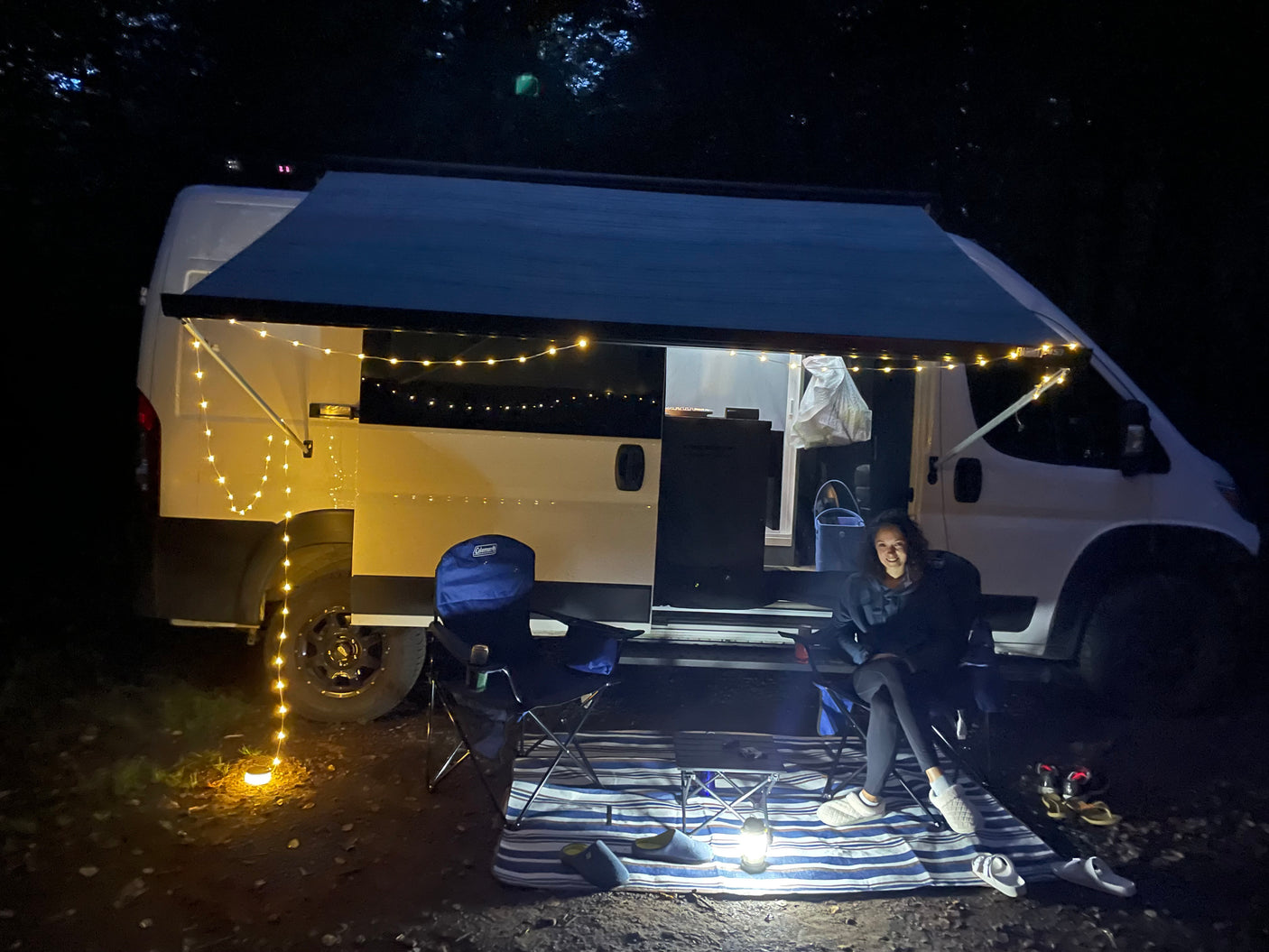A person relaxes by their white RV rental camper van at night. String lights illuminate the awning, creating a cozy outdoor camping scene.