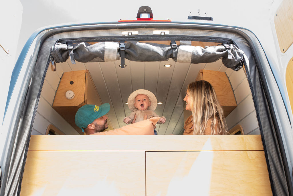 An expressive baby in a hat with parents in an RV rental, looking out their camper van's back door. Ideal for family travel.