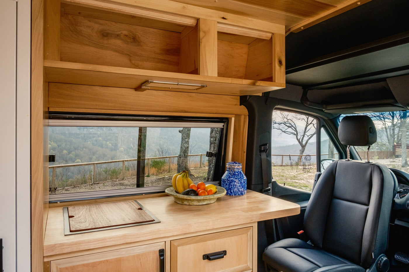 RV rental: light wood kitchen with fruit, overhead storage, and a scenic window view of nature from inside the van. — photo 14