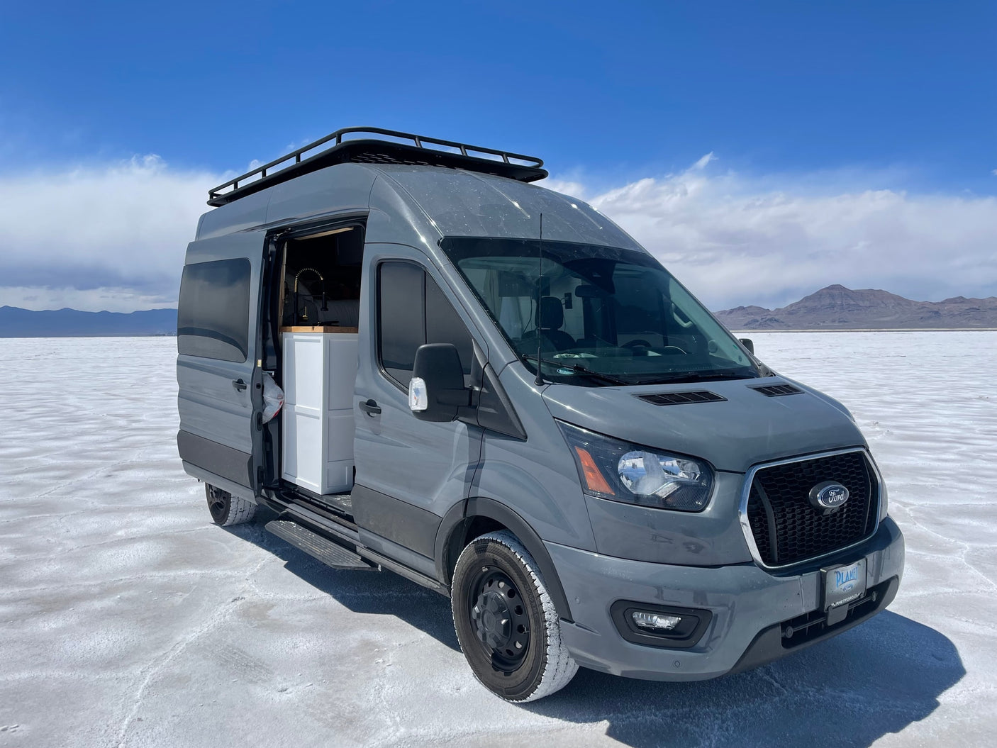 Grey Ford Transit camper van RV rental parked on vast white salt flats under a blue sky, ideal for adventures.