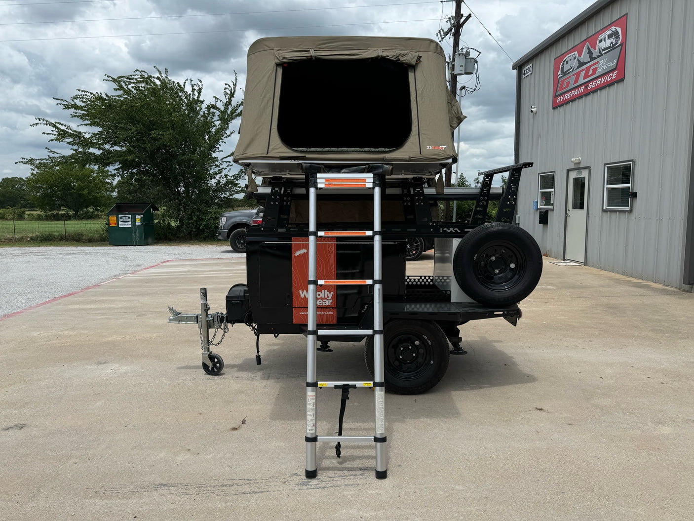 A camper trailer with an open rooftop tent and ladder, parked outside an RV rental and repair service building.
