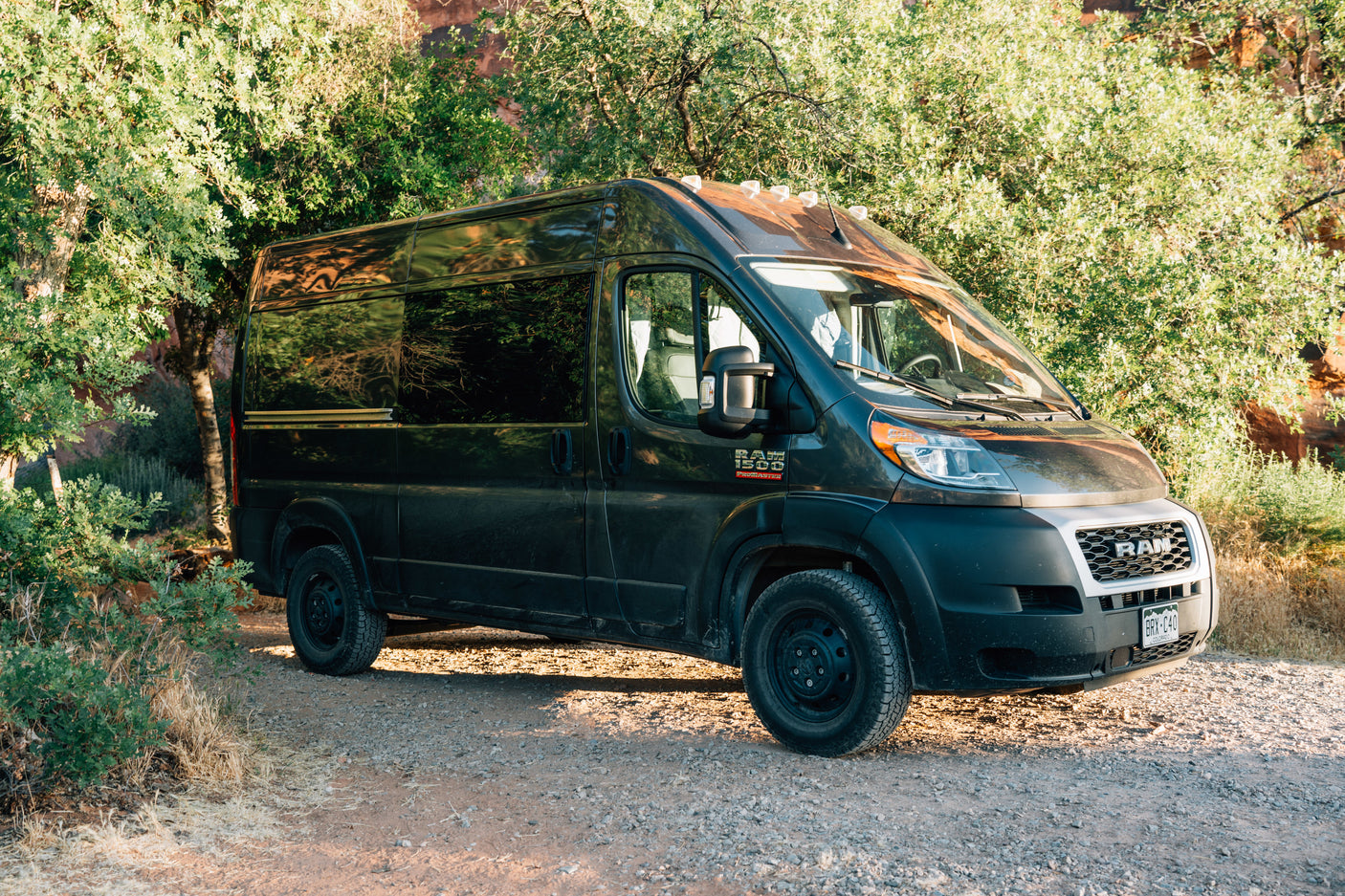 Experience nature with this black Ram ProMaster rv rental camper van, parked on a sunlit gravel road amidst green trees.